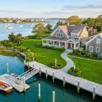 aerial view of a well-maintained Martha's Vineyard cedar-shingled coastal home with a clean dock in the foreground, clean stone pavers leading to the front door, lush green spring lawn